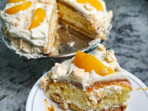 Whole cake with white frosting and candied peach slices atop, beside a plated slice revealing sponge and filling layers.