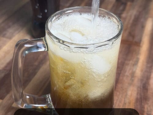 Clear liquid pouring into a glass mug filled with ice and pale soda, bottle behind on wooden surface